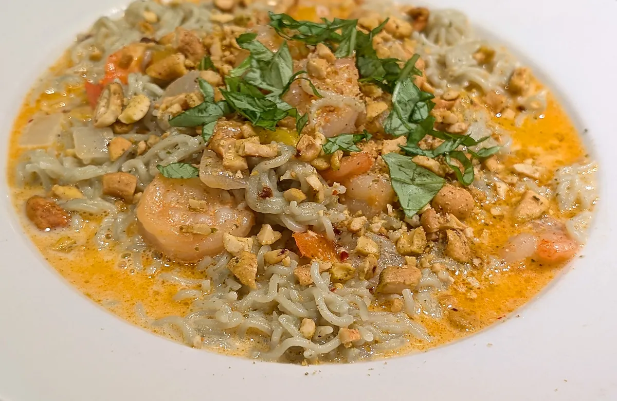 White rimmed bowl of shrimp and jade rice ramen noodles in a creamy orange coconut curry with bits of bell pepper and onion, topped with coarsely crushed cashews and thin ribbons of fresh basil